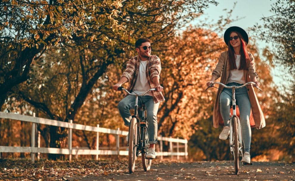A man and woman cycle along a country road, lined by trees covered in fall leaves.