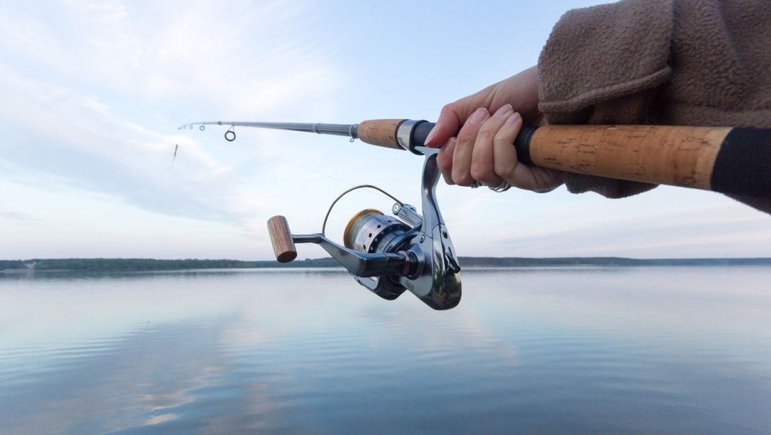Close-up of a person holding a fishing rod and reel over calm lake water.