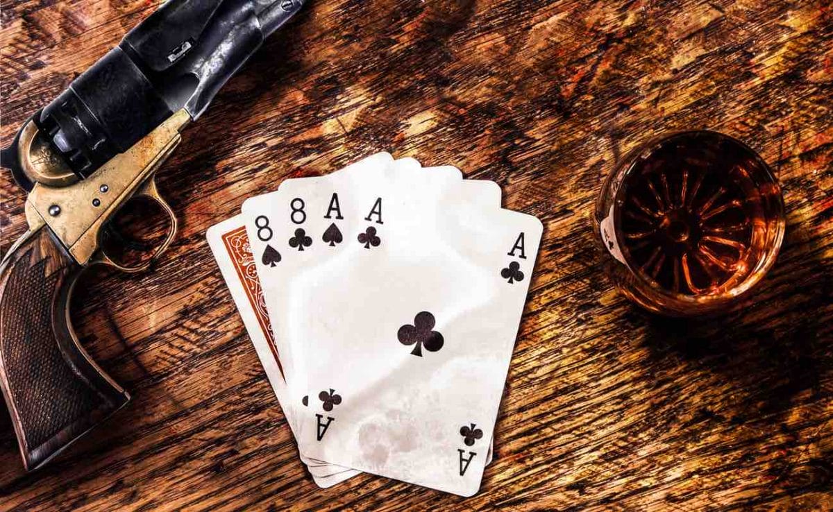 Top view of a table with playing cards showing a ‘dead man’s hand,’ a glass containing a drink and a colt revolver.