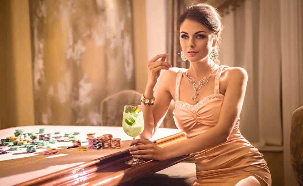 A woman in an evening gown holds a cocktail while sitting beside a casino table.