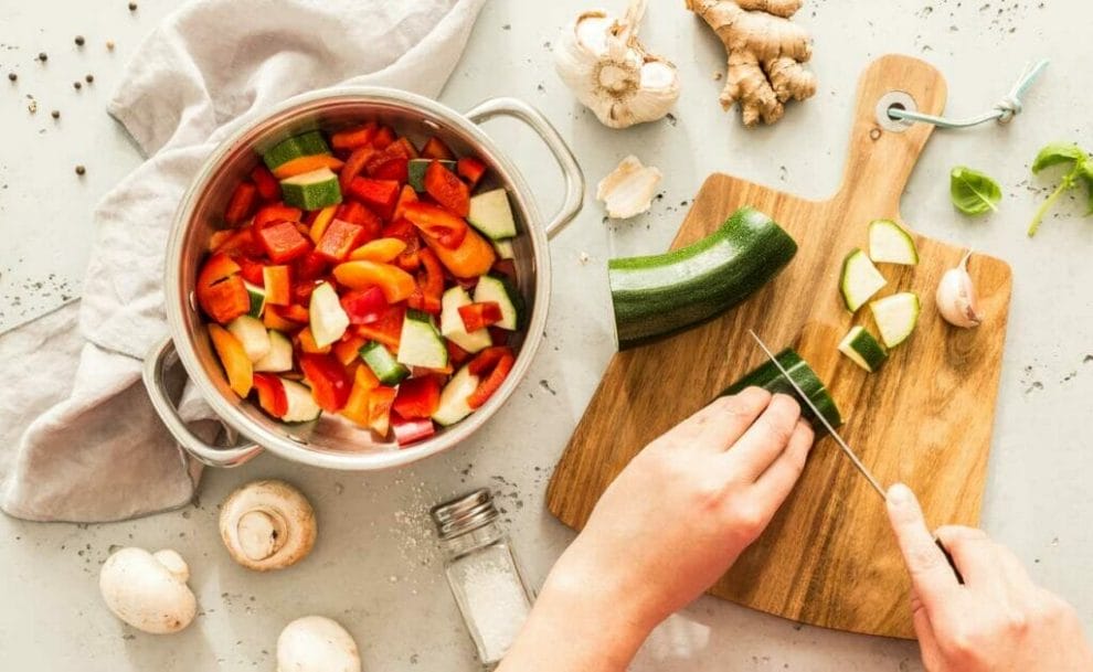 Hands chopping zucchini on a wooden board next to a pot of vegetables.