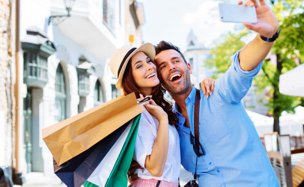 A man and woman with shopping bags take a selfie in the street outside a store.