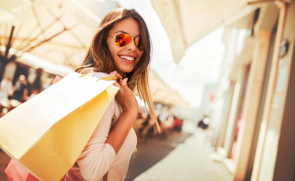 A smiling woman in sunglasses with shopping bags over her shoulder.