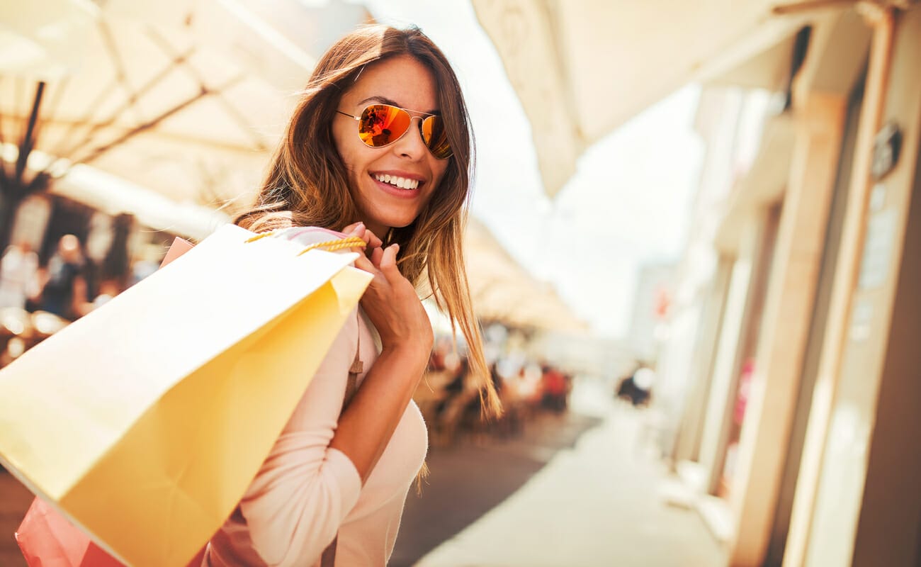 A smiling woman in sunglasses with shopping bags over her shoulder.
