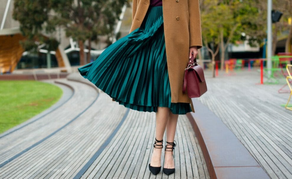 A bottom half picture of a woman wearing a green pleated skirt and a brown jacket.