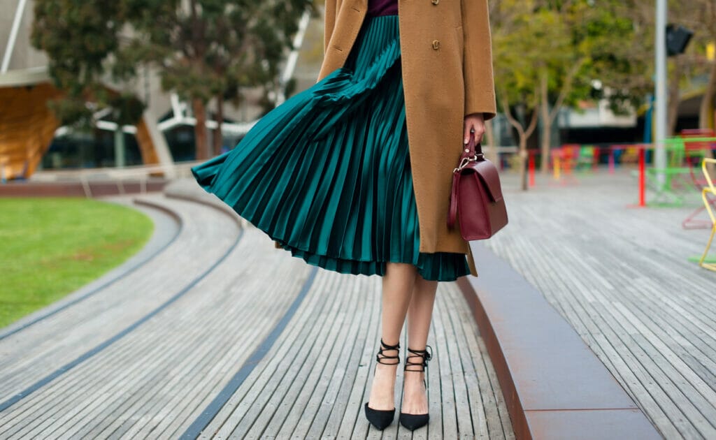 A bottom half picture of a woman wearing a green pleated skirt and a brown jacket.