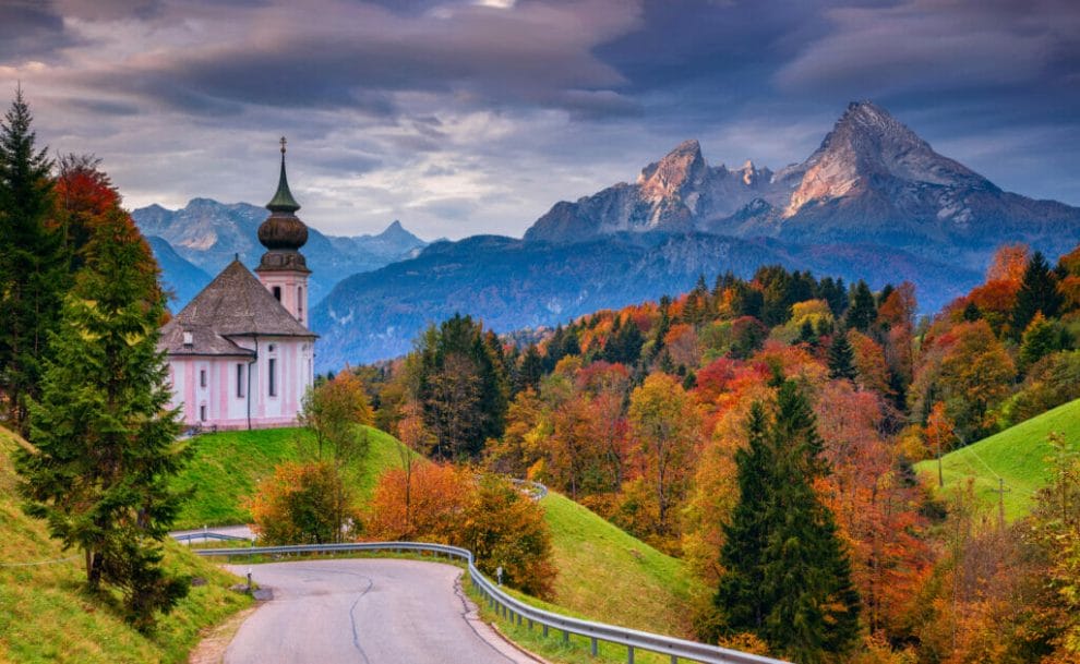 A road with a small church and trees with the Bavarian Alps in the background.