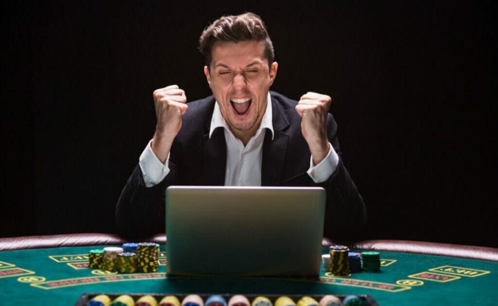 A man wearing a suit celebrates a win in front of his laptop. He is playing at a casino table, surrounded by poker chips.