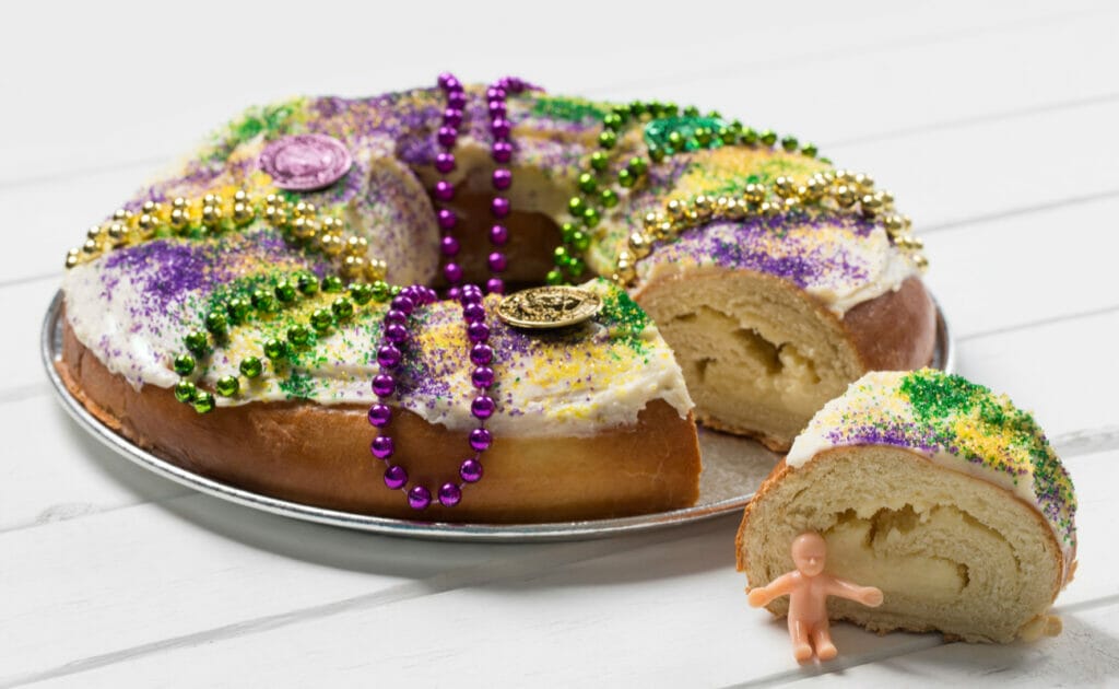 A decorated King cake with a plastic baby sitting next to a cut slice.