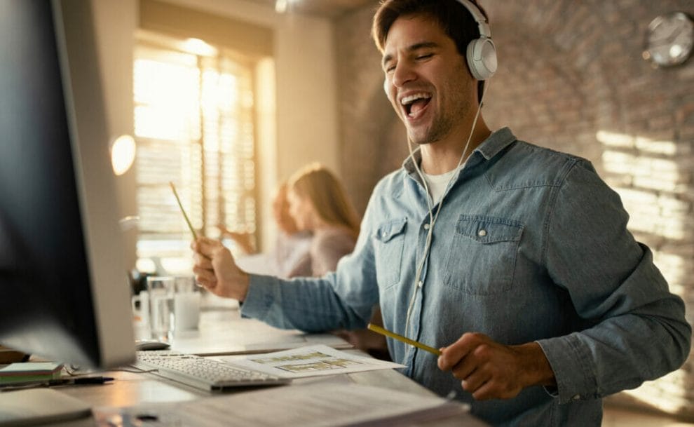 A young man listening to music at his computer and using pencils as drumsticks.