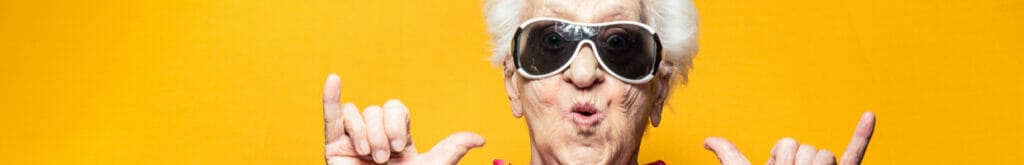 A funky older woman with her hands up and sunglasses on against a yellow background.
