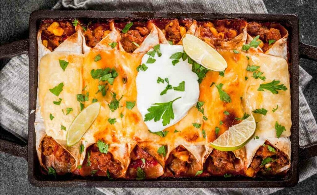 Top view of freshly baked enchiladas in an oven tray, topped with sour cream, cheese and lemon wedges.
