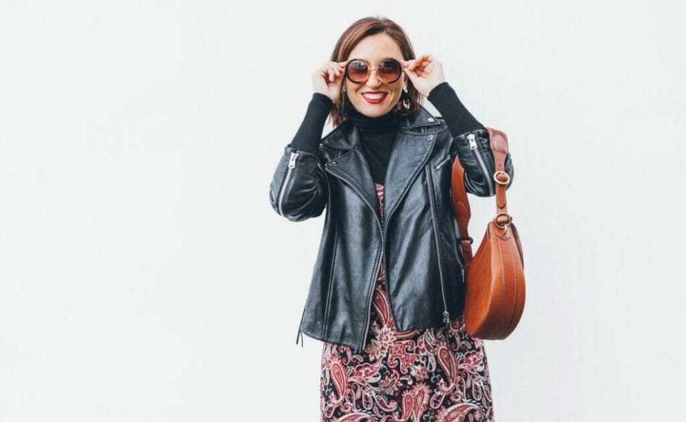 A smiling woman holding her sunglasses and wearing a black biker jacket with a brown leather bag.