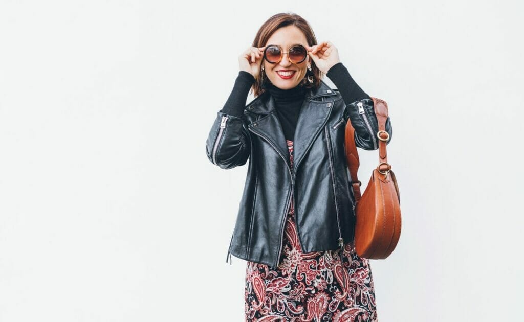 A smiling woman holding her sunglasses and wearing a black biker jacket with a brown leather bag.
