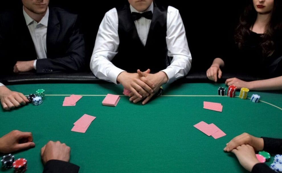 A dealer and four players sit around a green felt poker table. 