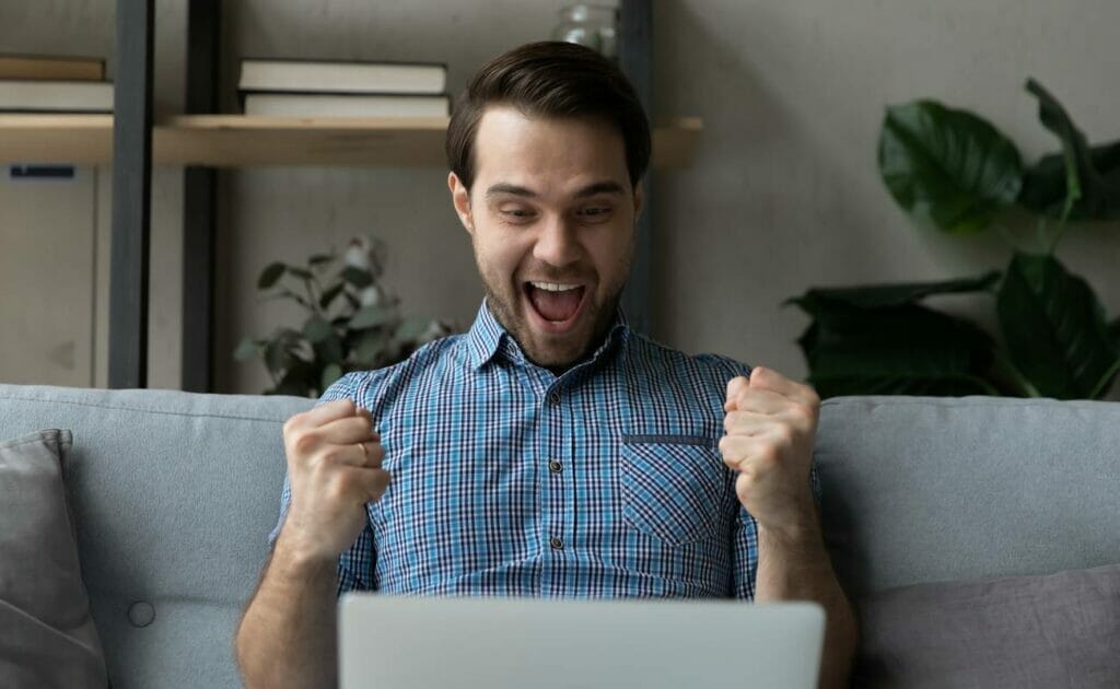 A man sitting on a couch with his laptop clenches his fists in excitement.