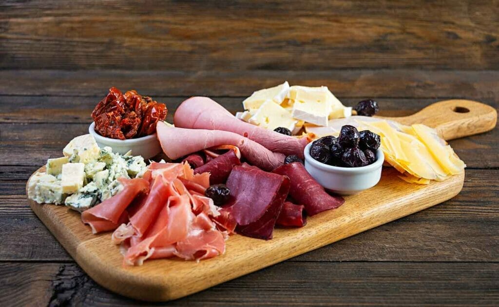 A snack platter on a wooden board with meats, cheese, and other food.