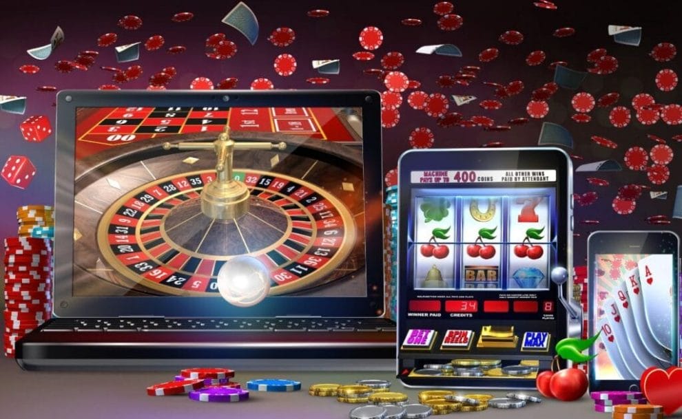 An online casino game concept image showing roulette on a laptop, slots on a tablet, and playing cards on a smartphone with poker chips, playing cards, and dice scattered around the background. 