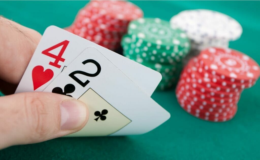 A player looks at a 4 of hearts and 2 of clubs in his hand with poker chips in the background