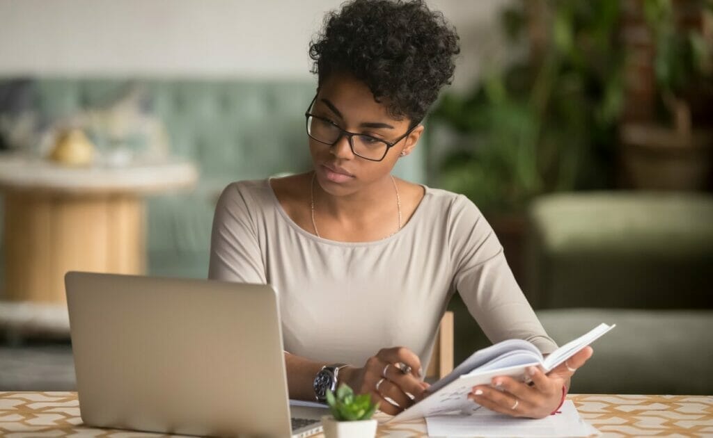 A woman wearing glasses looking at a laptop while holding a notebook.