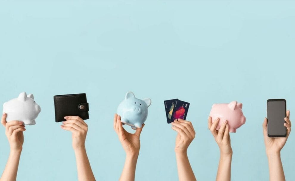 Hands holding up piggy banks, a wallet, bank cards and a cellphone.