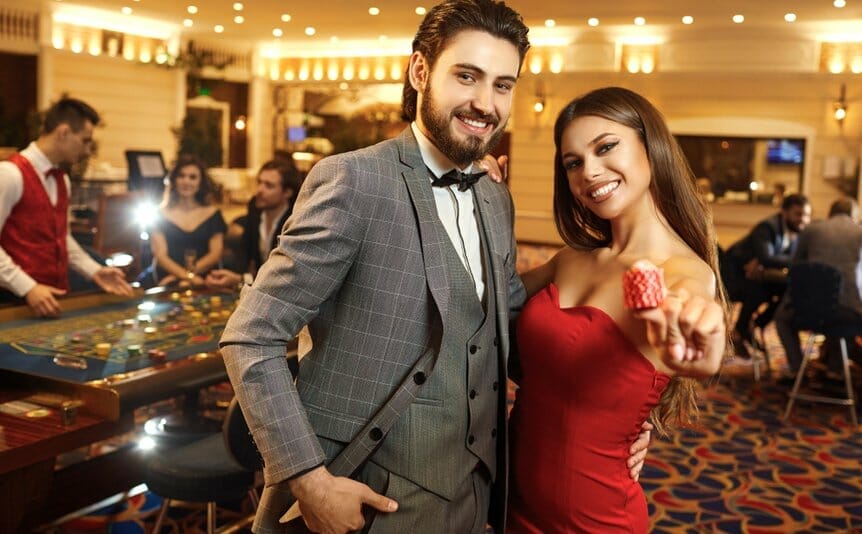A well-dressed couple poses in a busy casino while the woman holds a stack of casino chips.