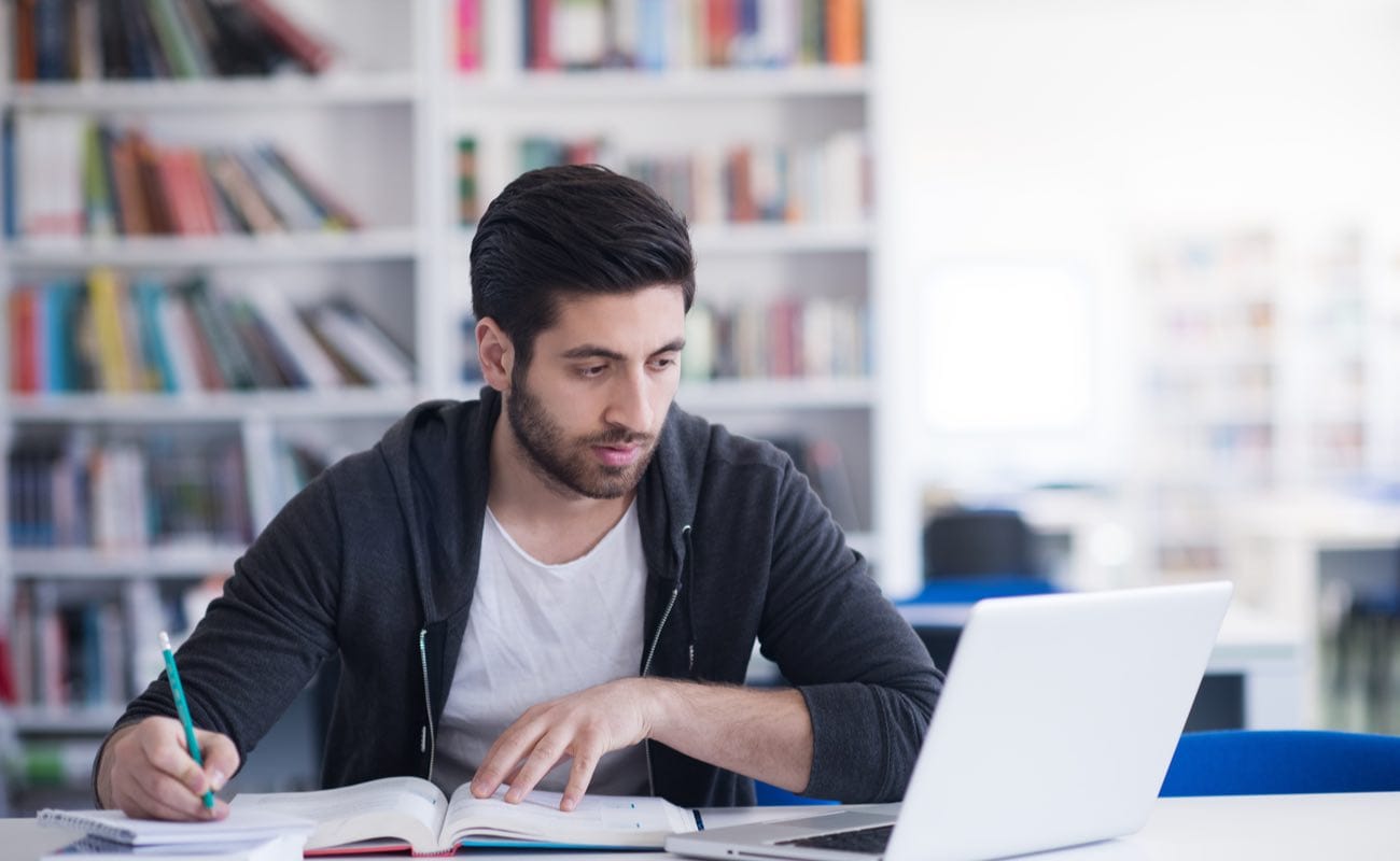 A man writing things down in a notepad while reading on a laptop.