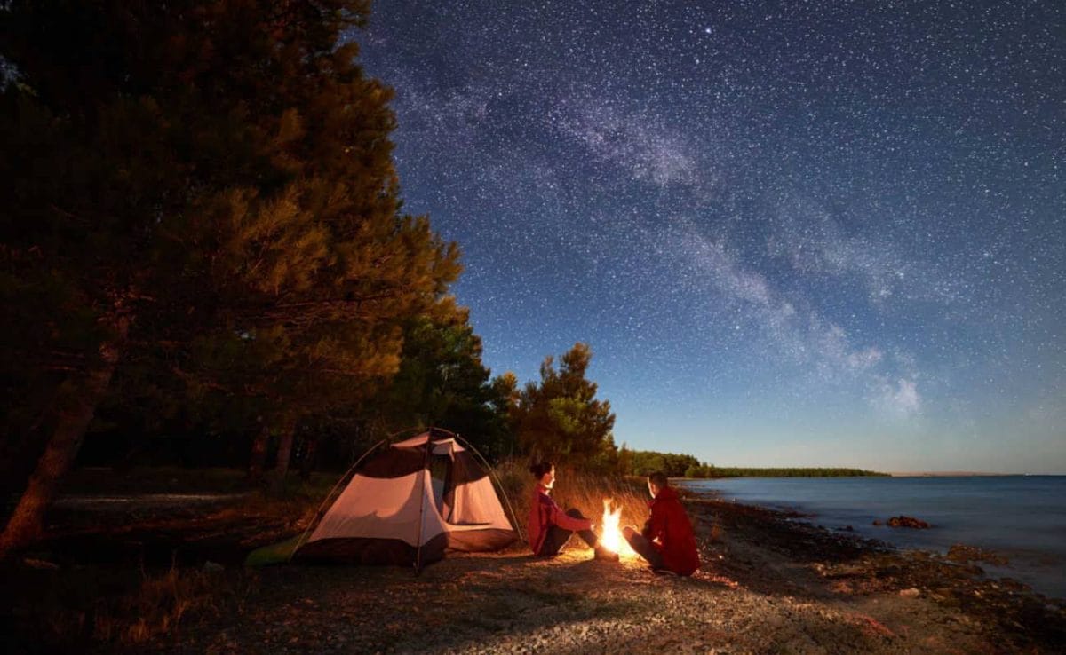 Two people sit around a campfire with a tent next to a lake at night.