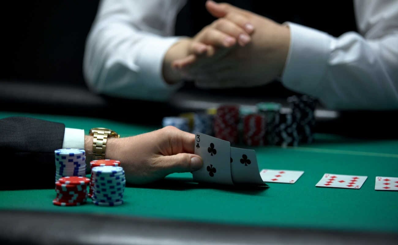 A man at a green poker table holding up two playing cards behind a stack of casino chips