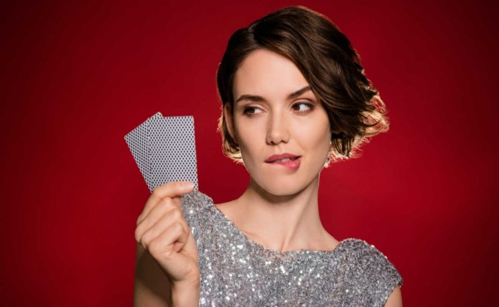 A woman biting her lower lip and holding up two playing cards against a red background.
