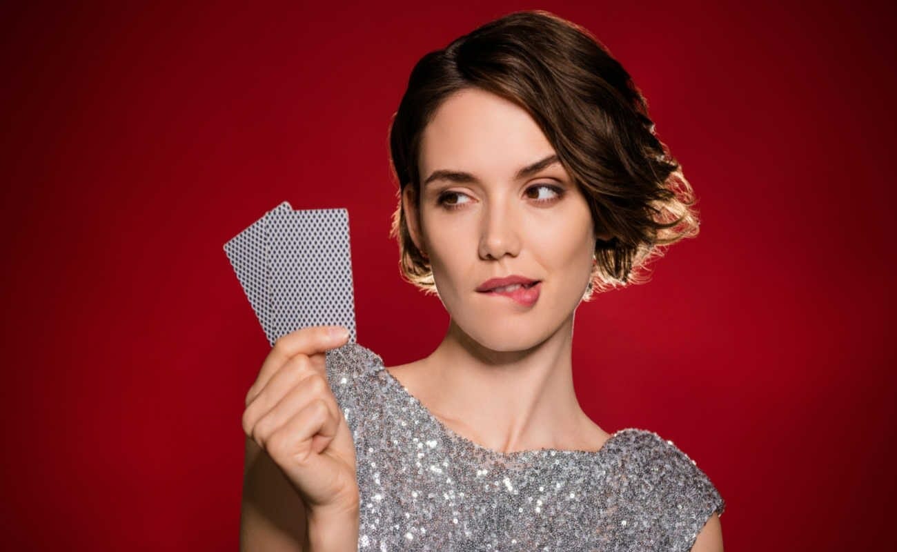A woman biting her lower lip and holding up two playing cards against a red background.