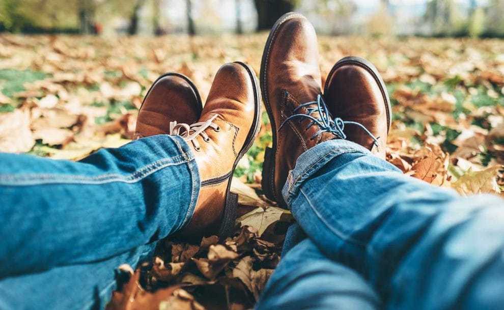 A man and woman sit in the autumn leaves in their jeans and boots.