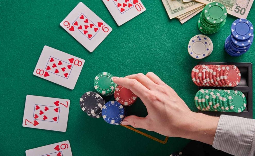 A hand reaching for poker chips on a green felt table with money and playing cards.