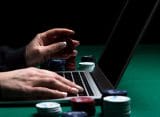 A person playing on a laptop with some poker chips next to him.