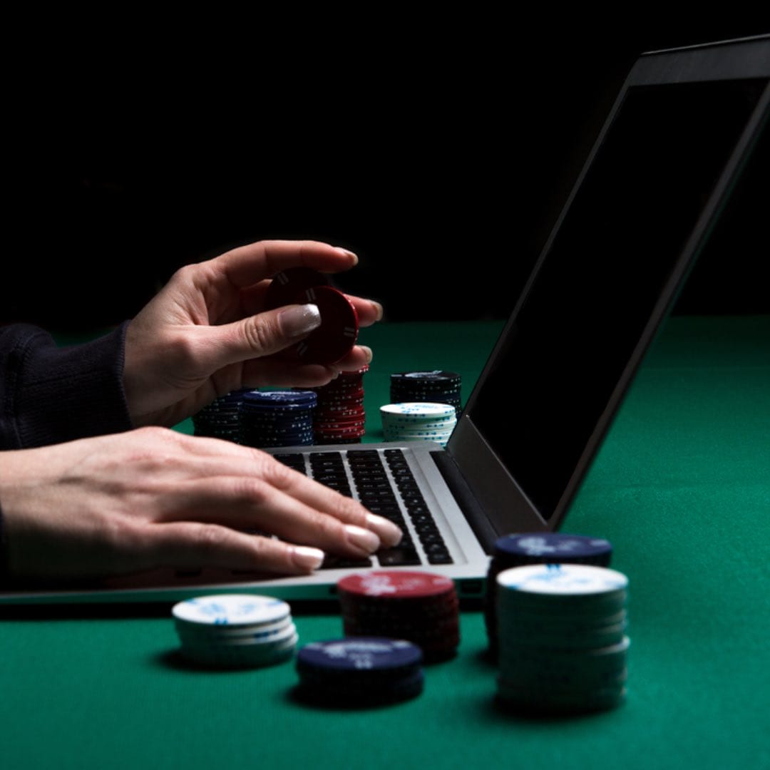A person playing on a laptop with some poker chips next to him.