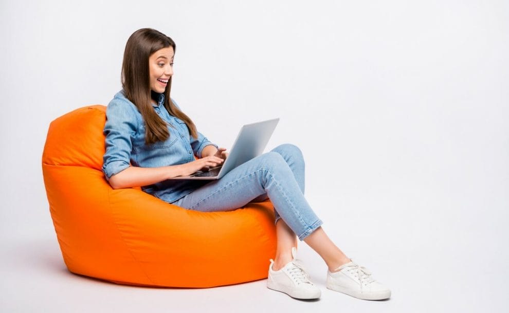 A woman smiling while sitting on a beanbag and using her laptop.
