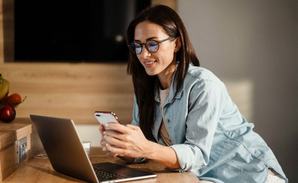 A woman looking at a smartphone with a laptop on the table in the background.