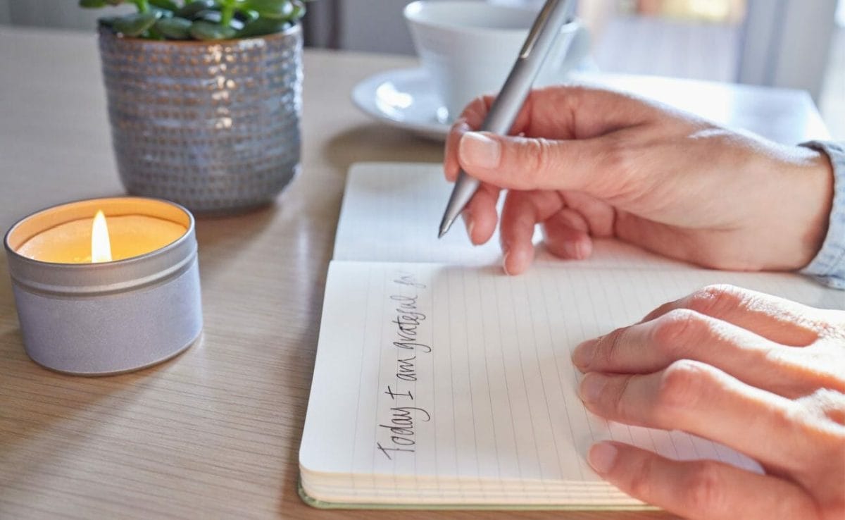 A person writing in a gratitude journal at a table with a candle and plant