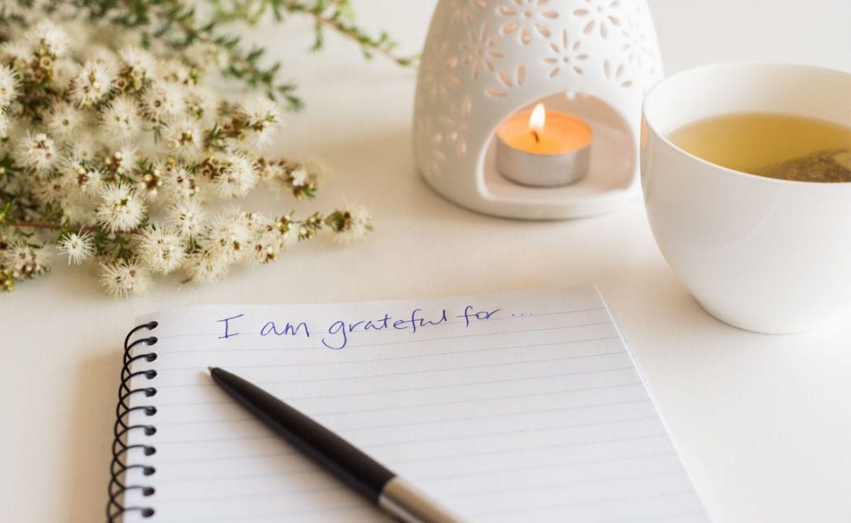A gratitude journal next to flowers, a pen, a tea candle and a cup of tea.