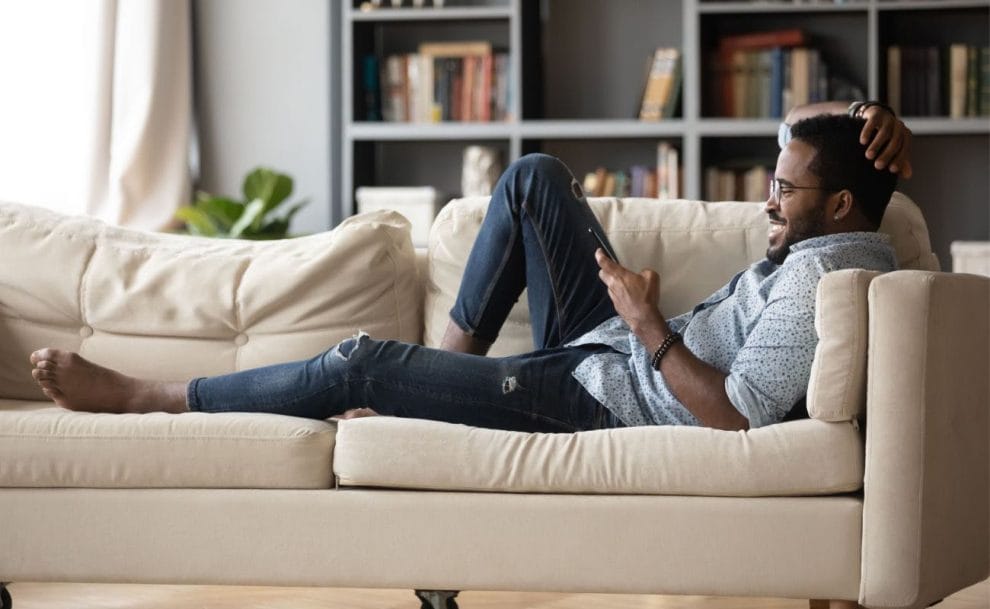 A man relaxing on a couch is smiling while looking at his smartphone.