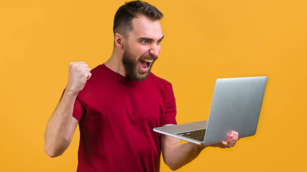 A person against an orange background celebrates while looking at their laptop 
