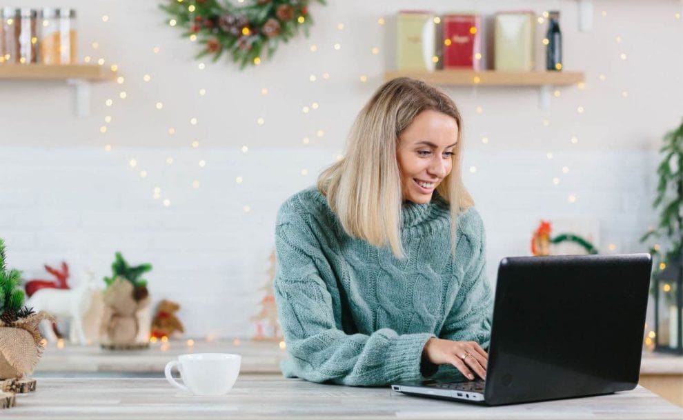  A smiling woman surrounded by Christmas decorations looks at her laptop screen.