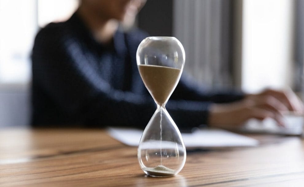 An hourglass on a wooden table with a woman in the background.