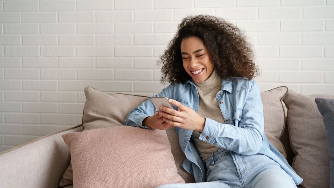 A happy woman looking at her smartphone while sitting on a couch.