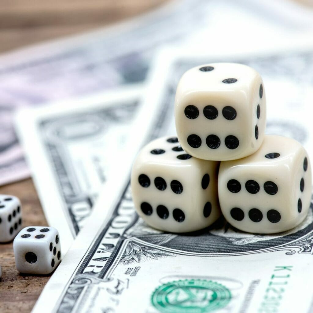 Three dice stacked on top of dollar bills on a wooden table, and two smaller dice beside the bills.