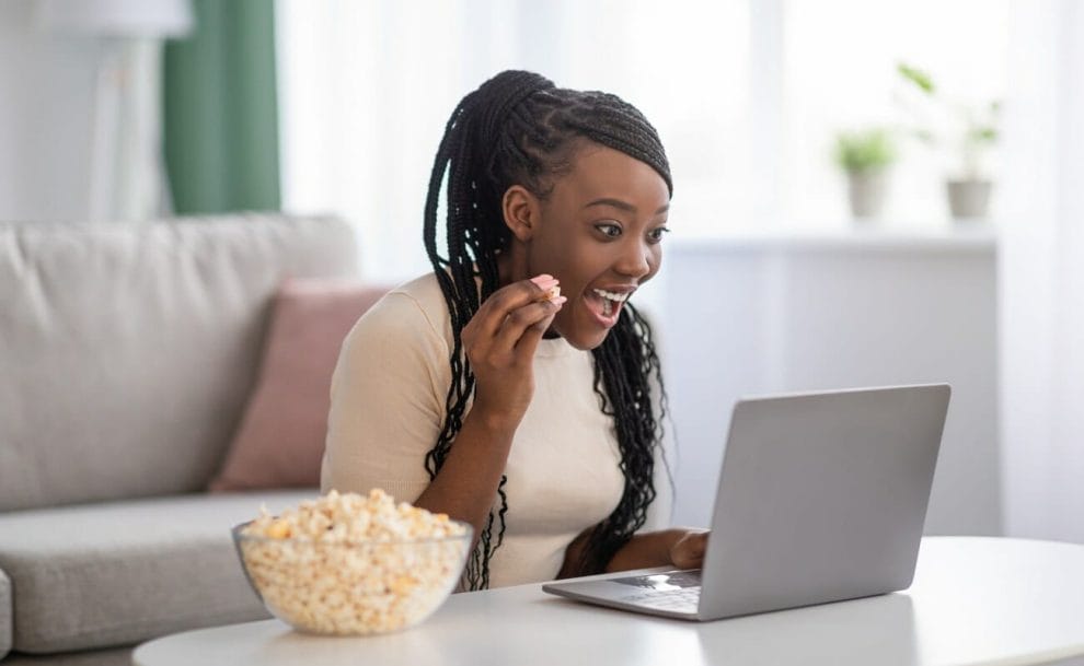 A young woman at a table with her laptop, smiling and eating popcorn.