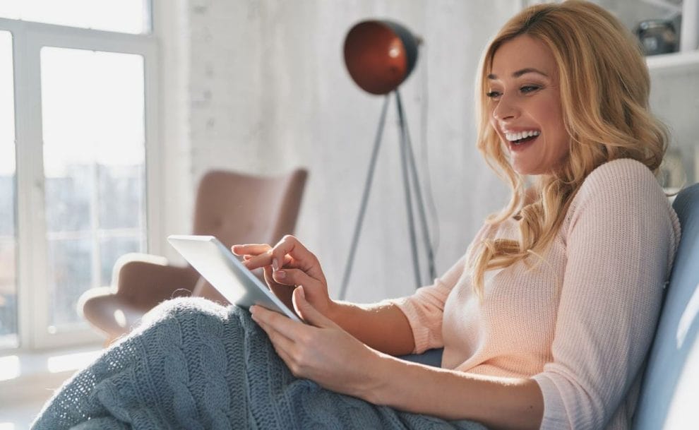 A smiling woman sits on a couch using a tablet.
