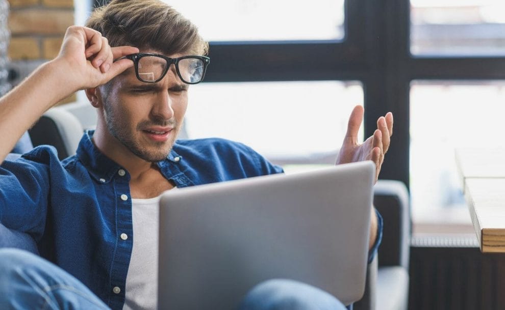  A young man using a laptop looking confused and lifting up his glasses.