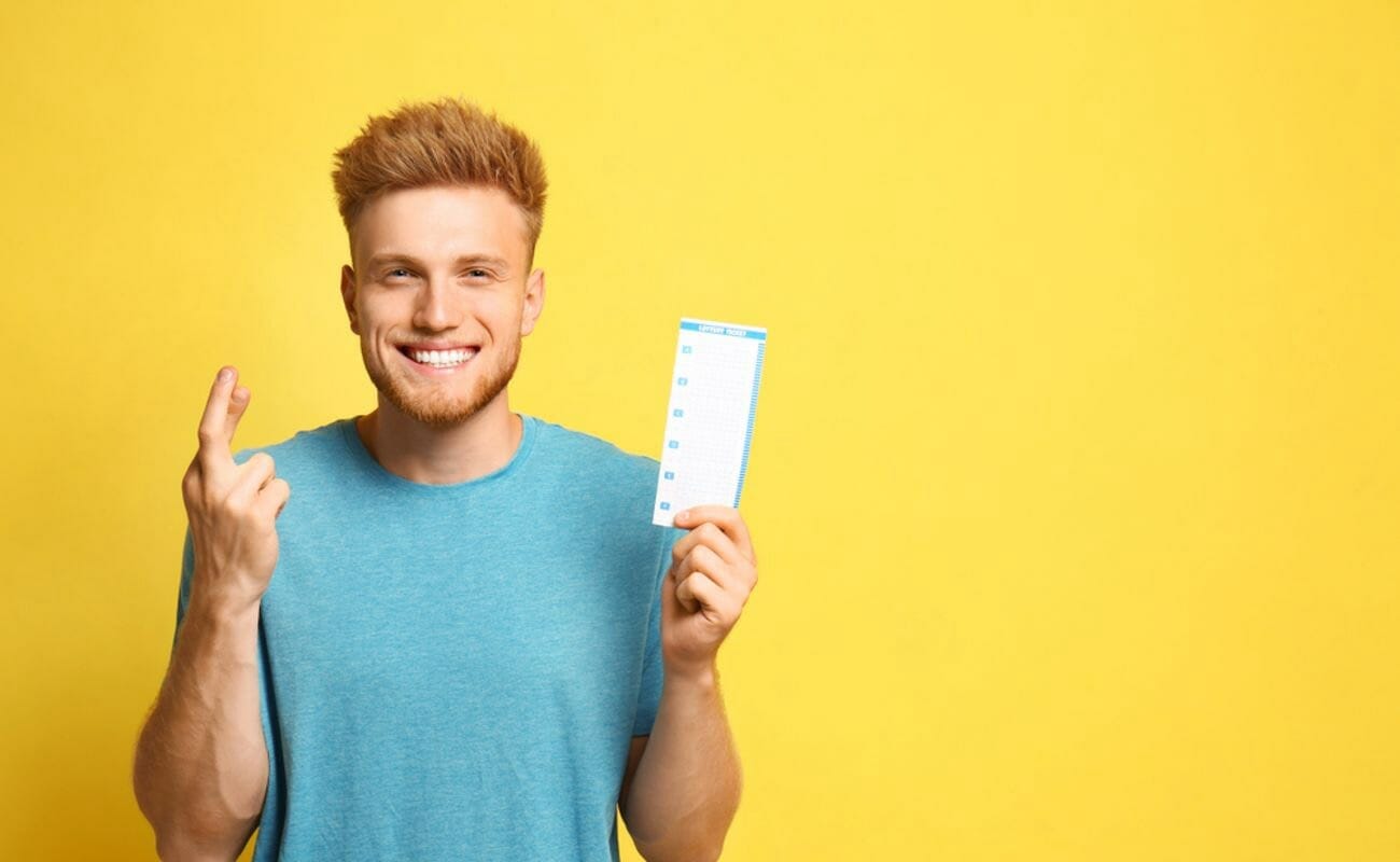 A smiling person against a yellow backdrop holds up a lotto ticket while crossing their fingers.