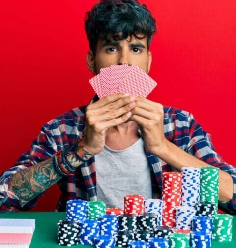 A poker player in front of a red wall and piles of poker chips holds his cards over his mouth.